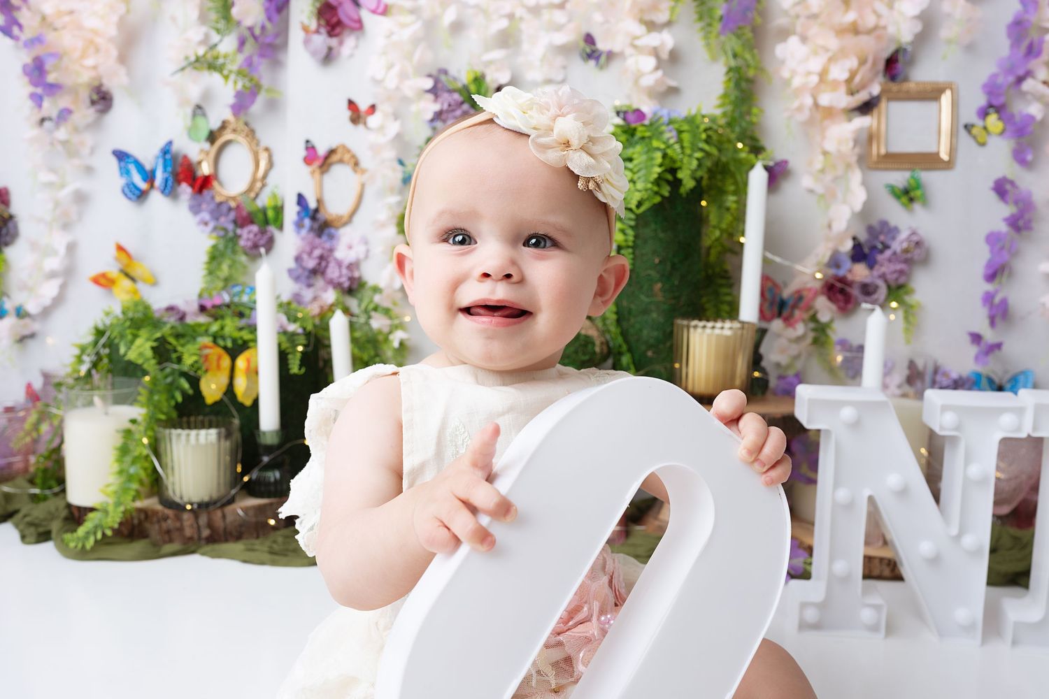 Baby in white dress sits with decorative number against floral backdrop with candles.