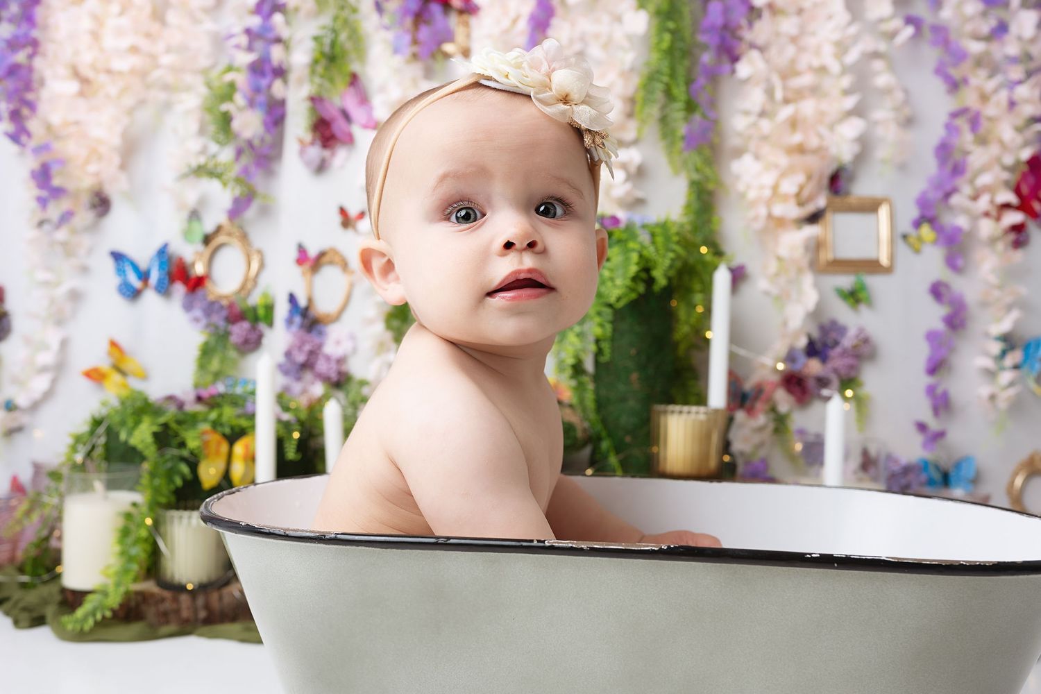Baby sits in vintage white enamel basin against floral backdrop.