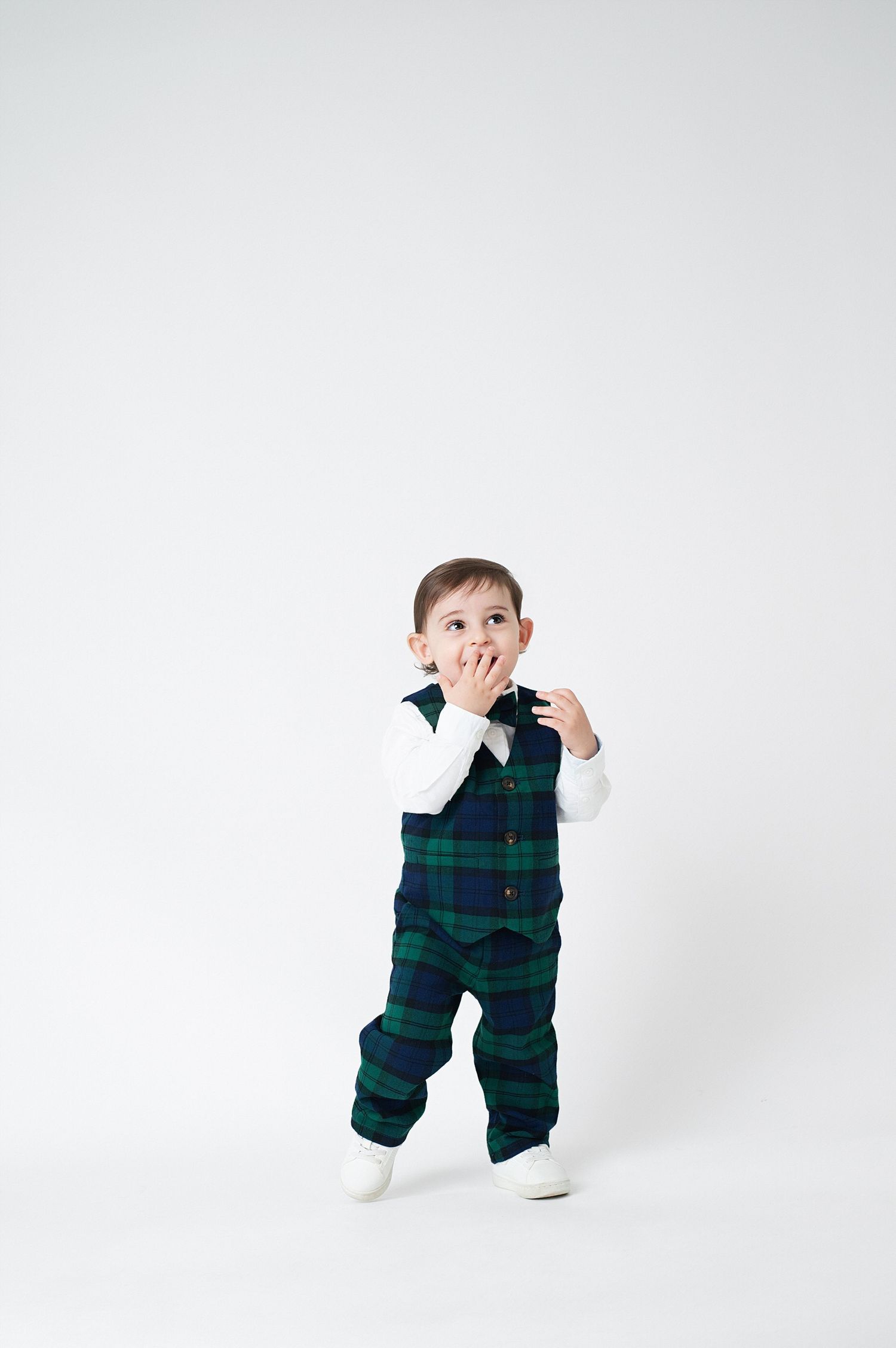 Toddler in plaid outfit covers mouth playfully against white background.