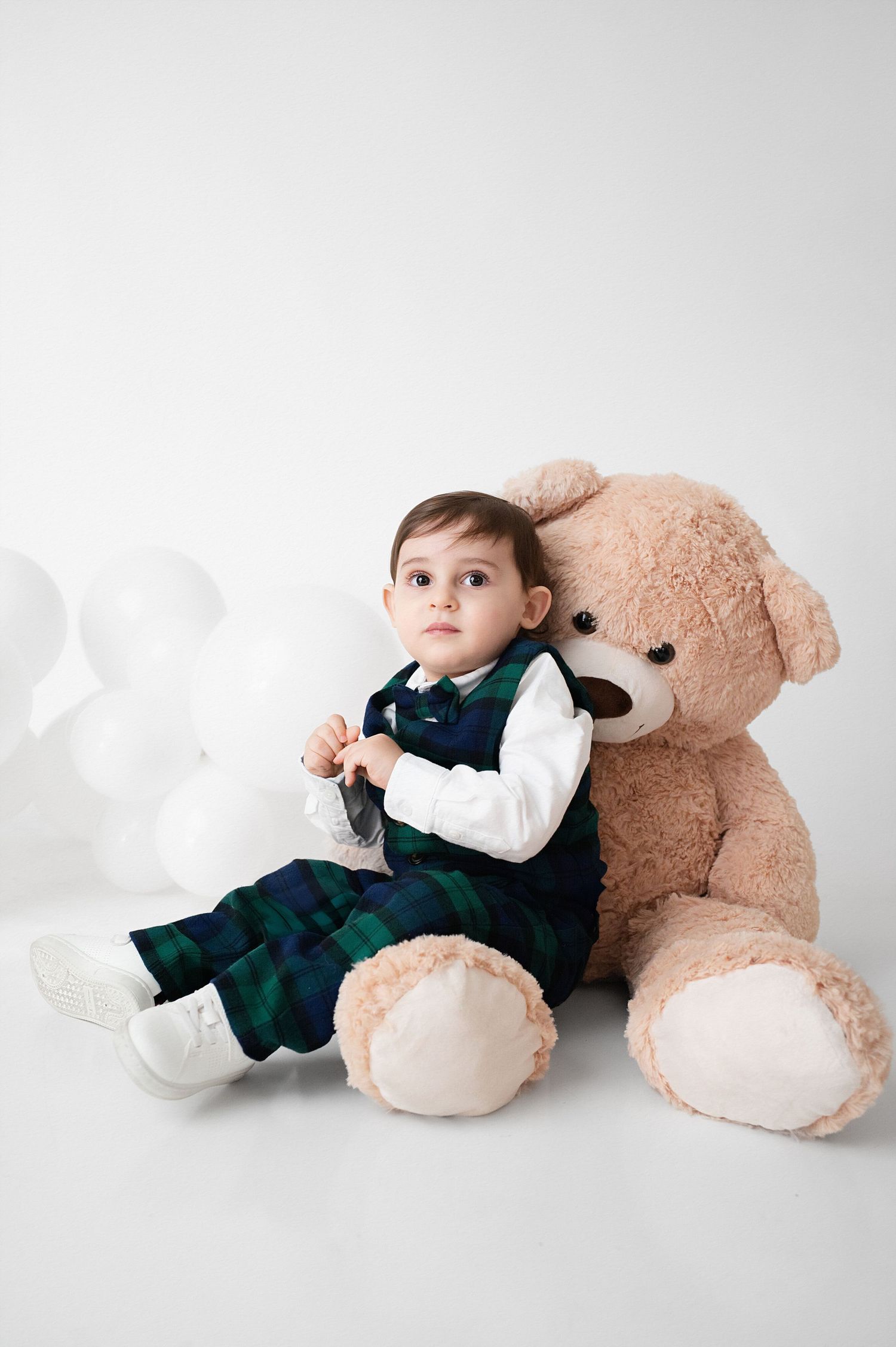 Young child in plaid outfit sits with large teddy bear against white backdrop.