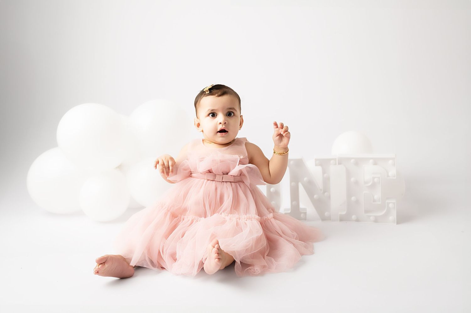 Baby in pink tulle dress sits by decorative letters against white backdrop.