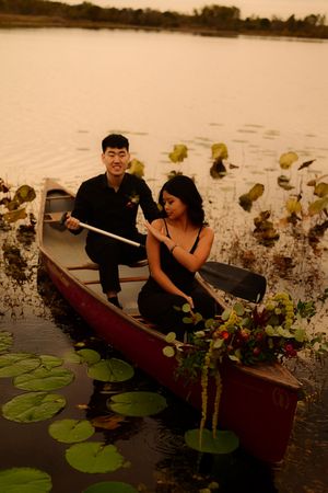 A couple in a wooden canoe explores a tranquil lake dotted with lily pads during sunset.