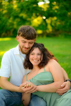 A couple embraces while sitting outdoors on grass with soft natural lighting and a green blurred background.