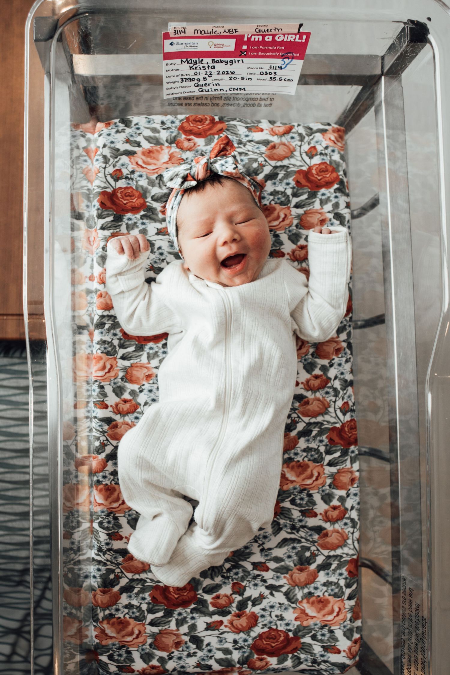 Baby in white outfit lying on floral blanket in hospital bassinet.