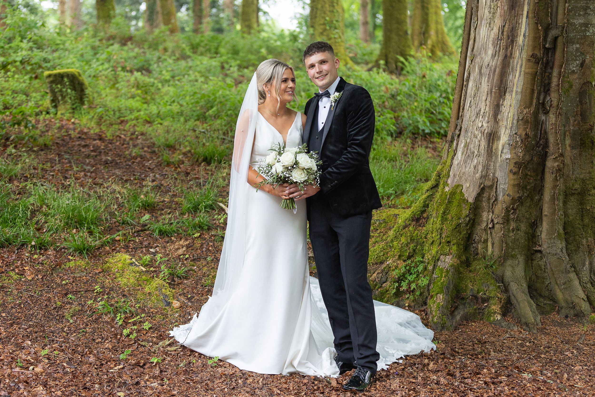 A wedding couple stands together in a lush forest setting with tall trees and mossy ground.  Dulrush Lodge Beleek County Fermanagh
