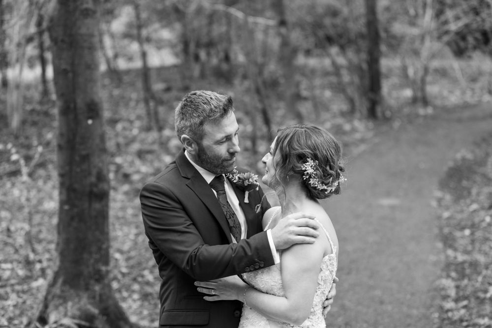 A romantic black and white photograph of a couple embracing on a wooded path during their wedding day.