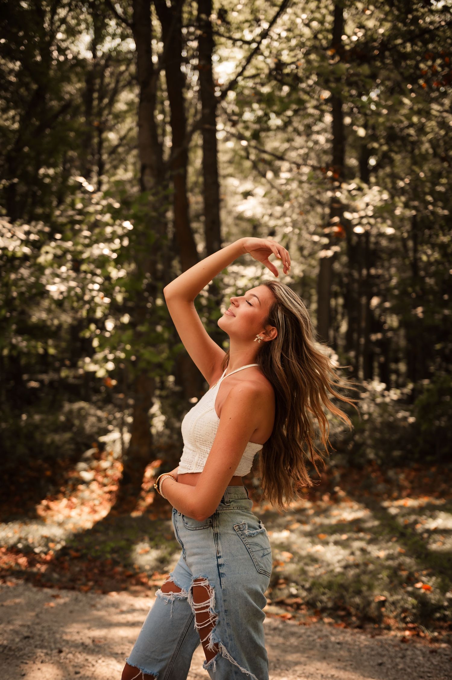 Young woman in ripped jeans tossing hair in sunlit forest setting.