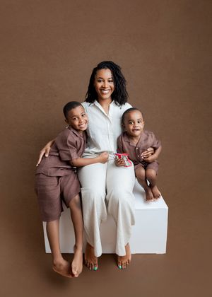 A smiling woman in white sits on a cube with two young boys in matching brown outfits against a brown backdrop.