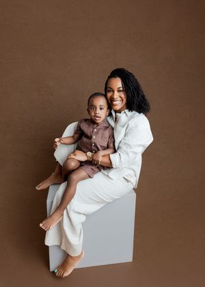 A smiling woman in white sits on a cube holding a toddler boy in a brown shirt against a brown backdrop.