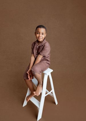 Young boy in brown shorts set sits on white stool, smiling at camera against warm brown studio background.