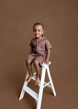 A smiling young boy in a brown linen outfit sits on a white ladder stool against a warm brown studio background.