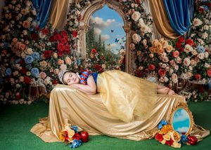 Girl dressed as Snow White in golden gown lying among colorful flowers in enchanted fairy tale themed photo.