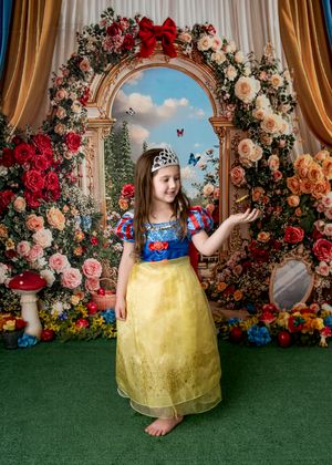 Young girl in Snow White costume with tiara poses before floral arch backdrop with roses and butterflies.