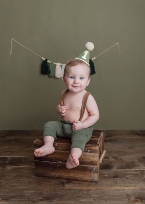 A baby in green overalls sits and smiles on a rustic wooden crate against a neutral wall.