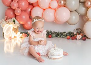 Baby sitting on floor with pink and rose gold balloon garland and birthday cake for first birthday photo session.