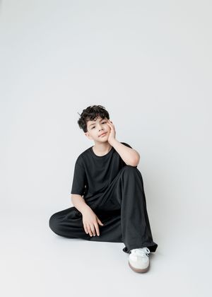 A young boy in a black outfit sits cross-legged on a white background, resting his cheek on his hand.