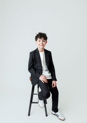 A smiling boy in a black blazer and white sneakers sits on a stool against a light gray background.