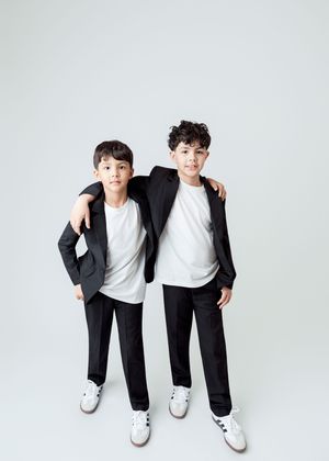 Two boys in matching black suits and white shirts pose together against a light gray background.