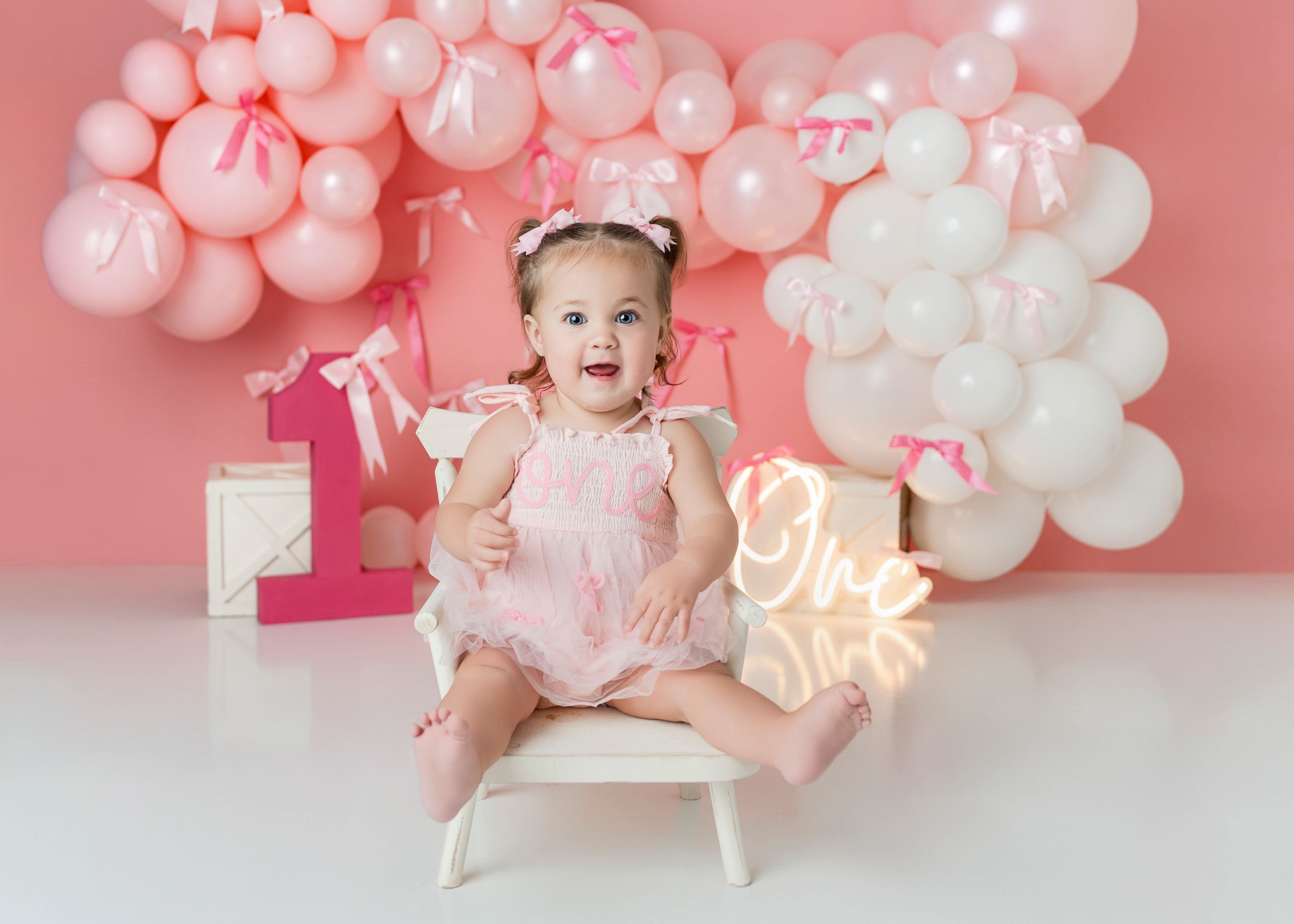 Pink and white balloon backdrop with party decorations and neon lighting for a first birthday photoshoot.