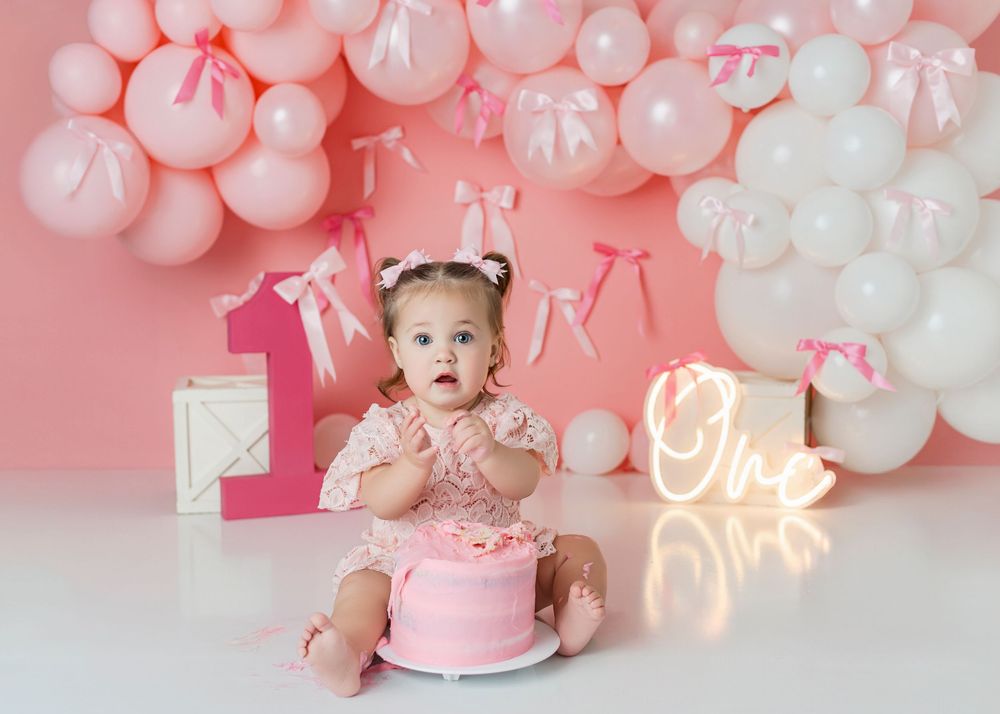 A pink and white balloon arch installation with neon signage creates a festive first birthday party backdrop.