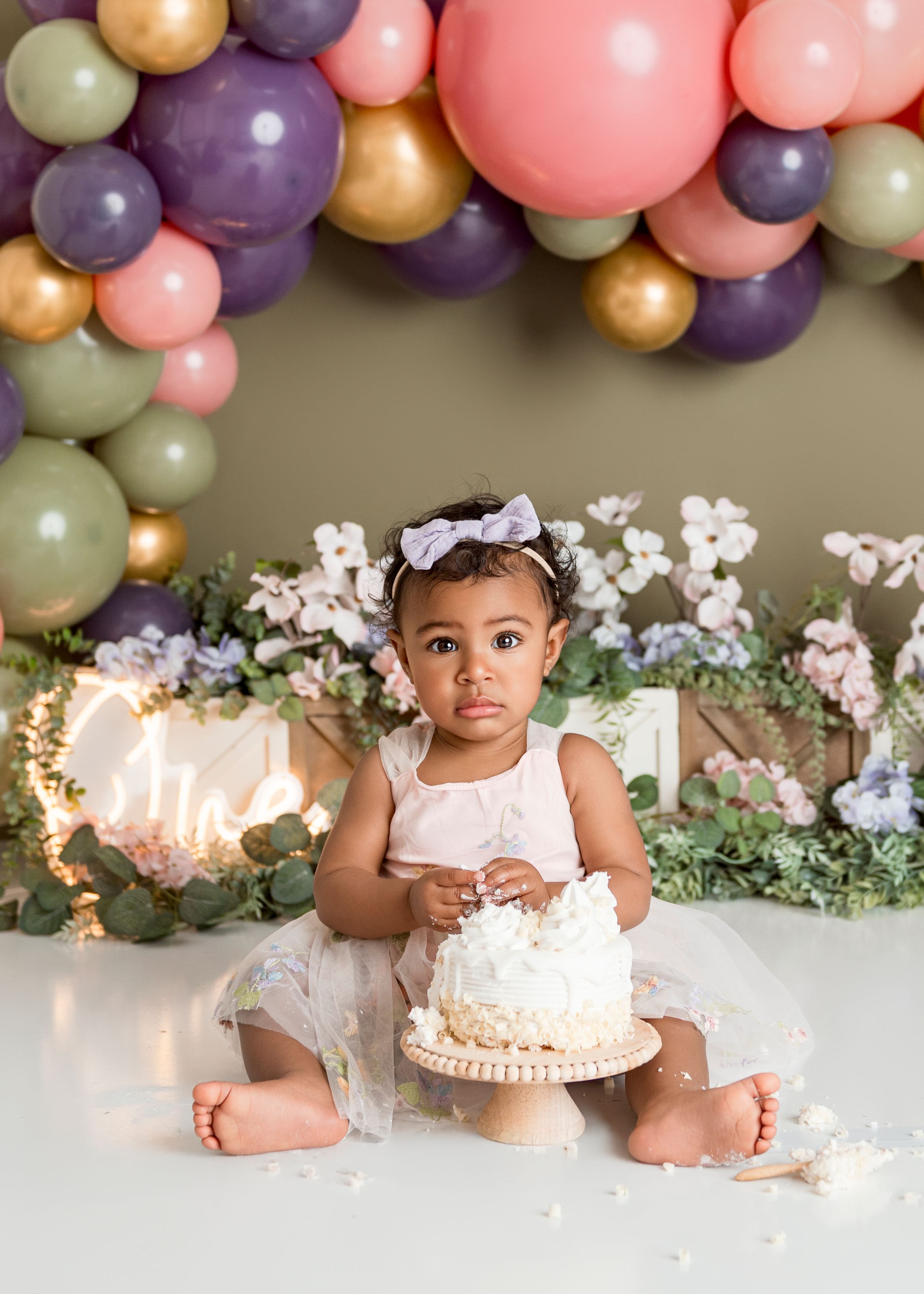 A baby in a white dress sits by a birthday cake under colorful balloon garlands and floral decorations.
