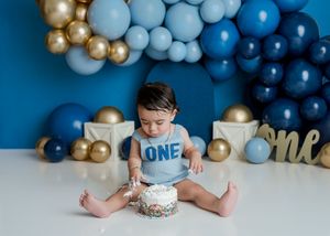 Baby in blue 'ONE' shirt sits with cake during first birthday photoshoot with blue and gold balloon arch decoration.