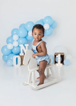 Baby boy in blue romper sits on white rocking horse, surrounded by blue balloon garland and ONE marquee letters.