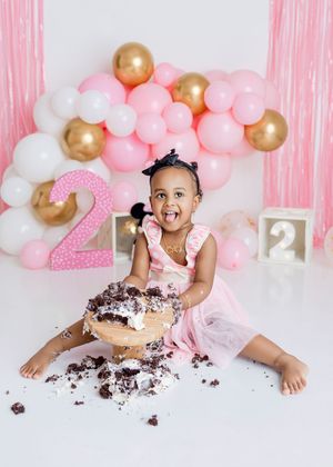 Toddler girl in pink dress smashing chocolate cake at 2nd birthday party with pink, white, and gold balloon backdrop.