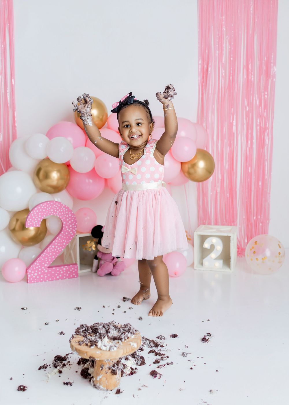 Toddler girl in pink tutu dress celebrates 2nd birthday smash cake with balloons and pink decorations.