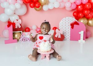 Baby girl in pink heart dress smashes first birthday cake surrounded by red, white, and gold balloon arch.
