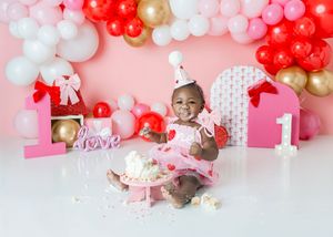 Baby girl in pink dress celebrates 1st birthday cake smash with red, pink, and gold balloon garland backdrop.