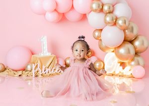 Baby girl in pink tulle dress celebrates first birthday with gold and pink balloon arch and number one cake.