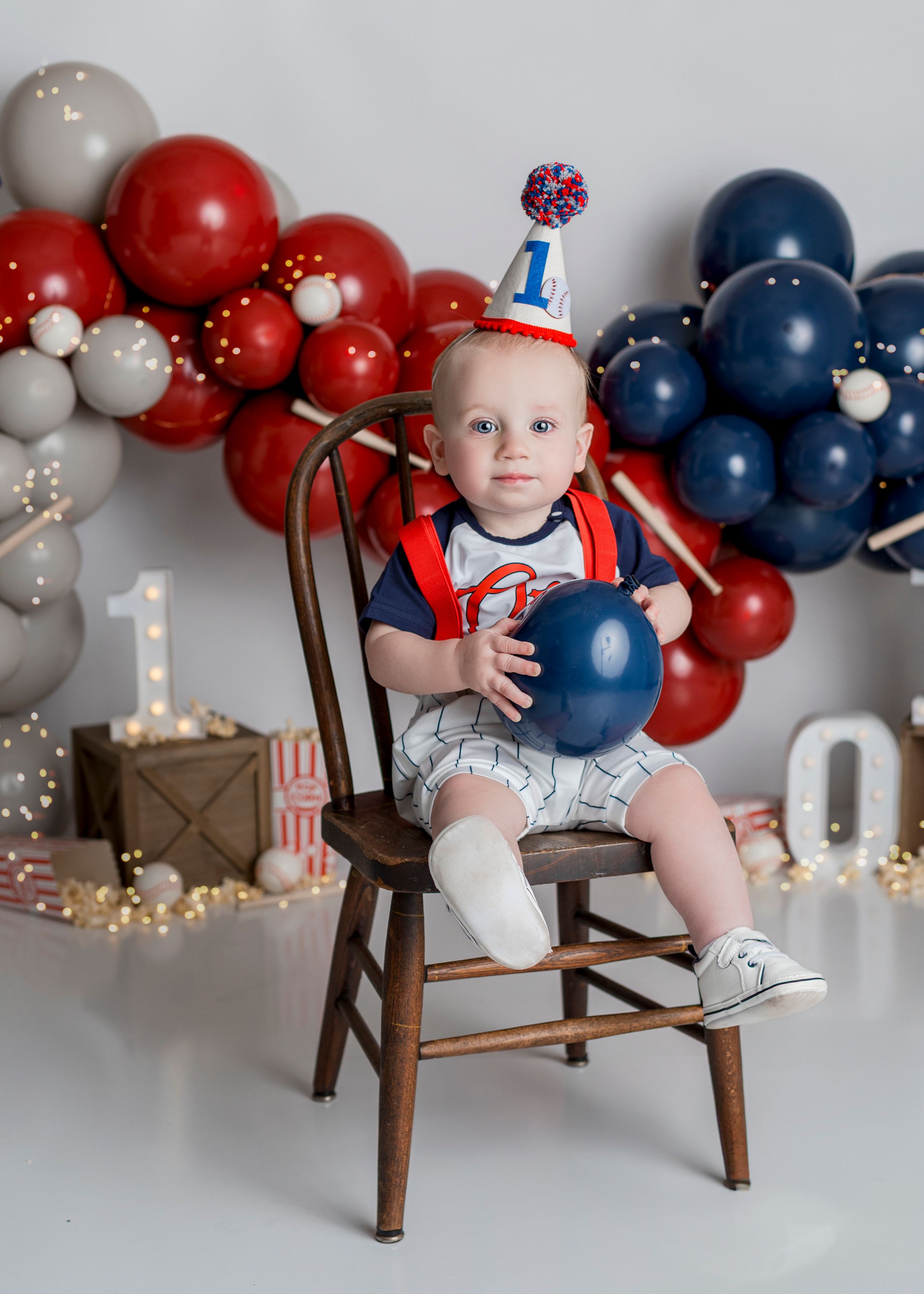 A baby in a baseball themed outfit sits on a wooden chair with red, white and blue balloon decorations in the background.