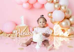 Baby girl in pink dress sits by first birthday cake with princess sign, pink and gold balloon arch on pink backdrop.