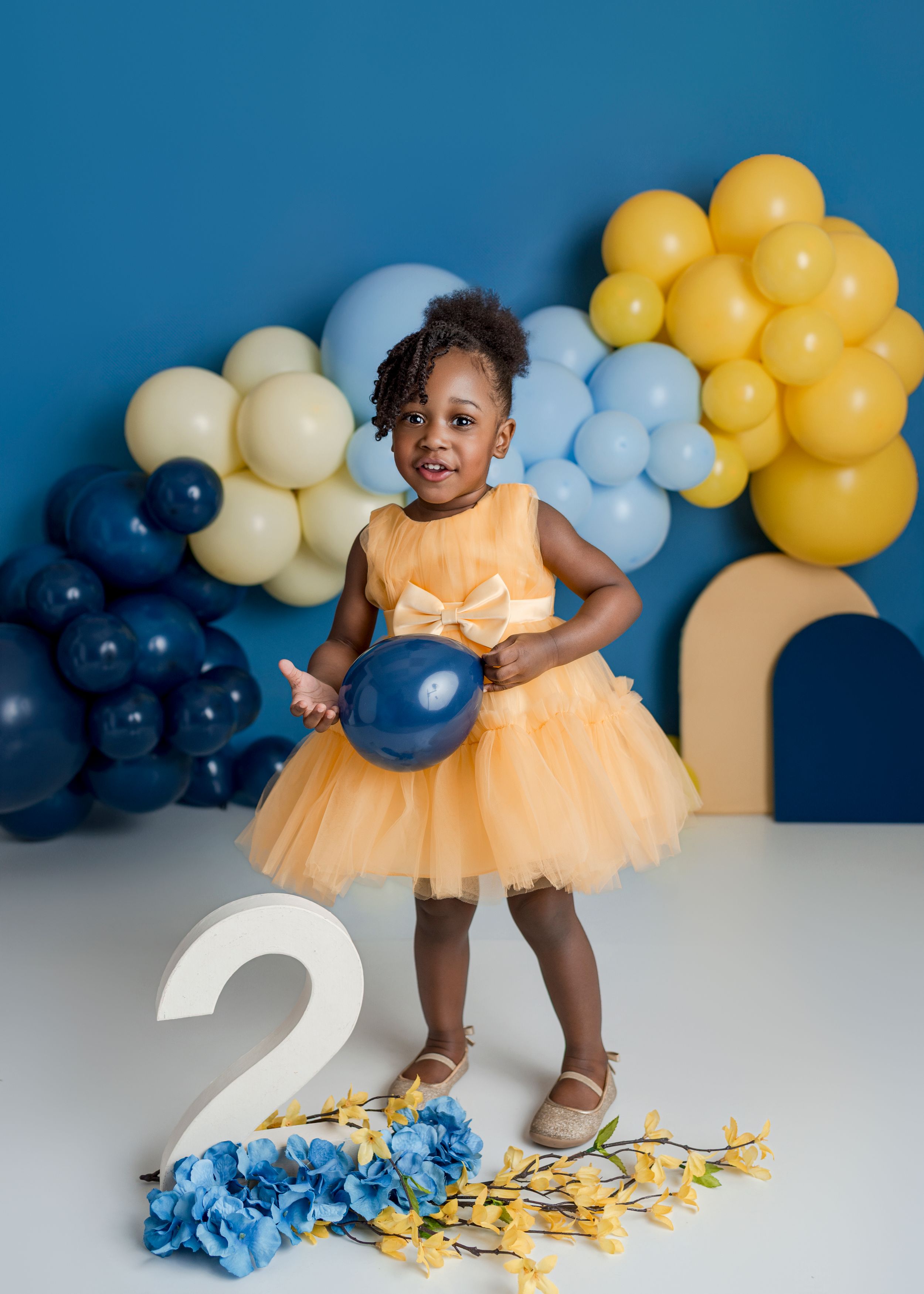 A festive birthday setup with navy, yellow and light blue balloons against a white backdrop features a large number 2.