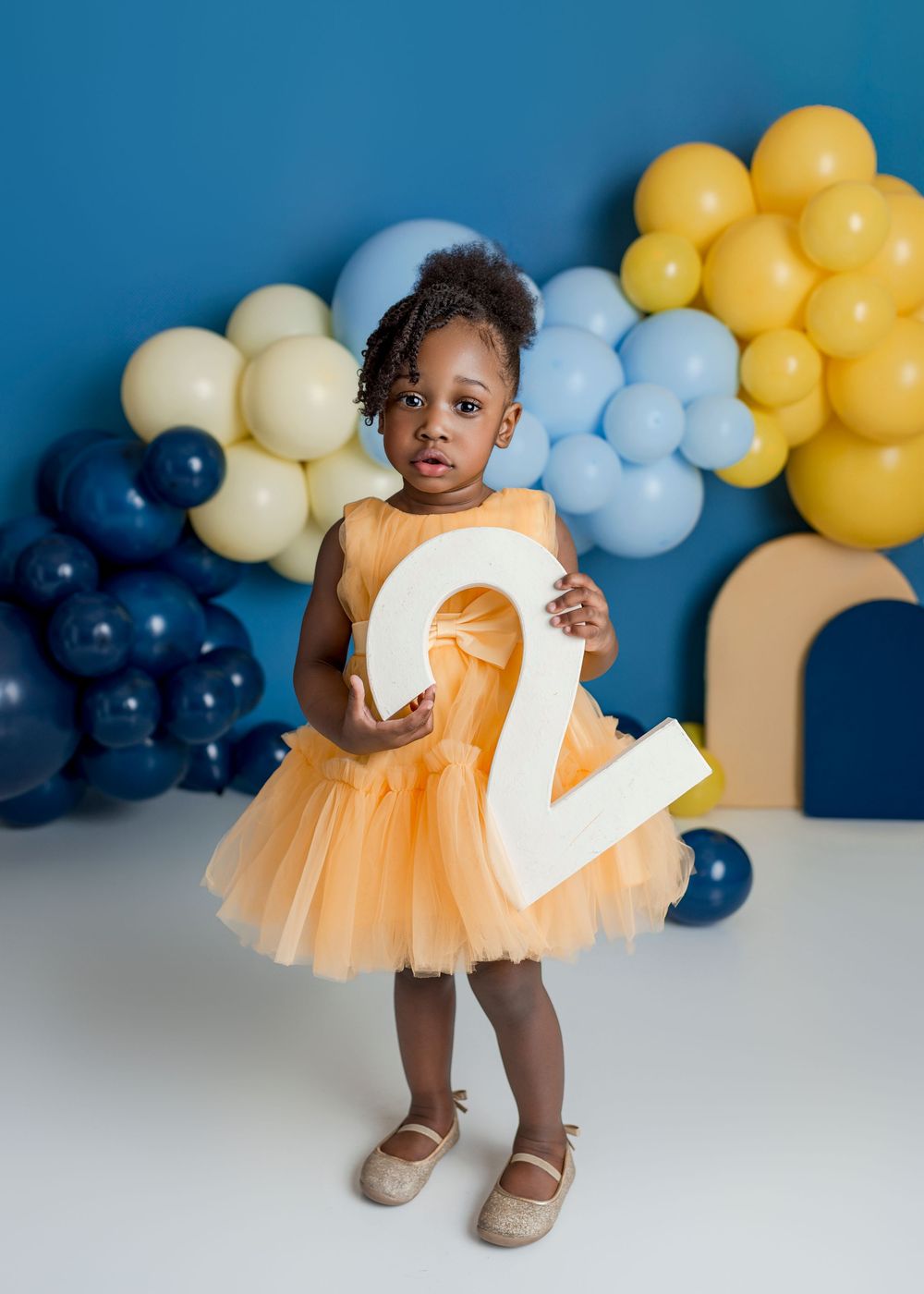 A young child in an orange tulle dress holds a large number 2 against a backdrop of blue and yellow balloons.