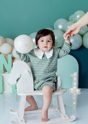 Baby in blue patterned romper sits on white rocking horse chair, surrounded by pastel balloons for first birthday.