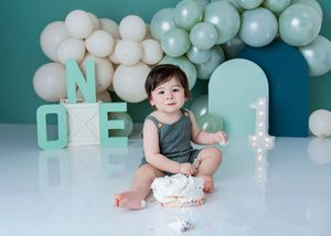 Baby's first birthday cake smash photo with teal ONE letters, white balloons, and adorable infant in green romper.