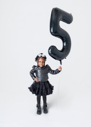 A young Black girl in a black leather jacket and tutu skirt holds a large black number 5 balloon, celebrating her birthday.