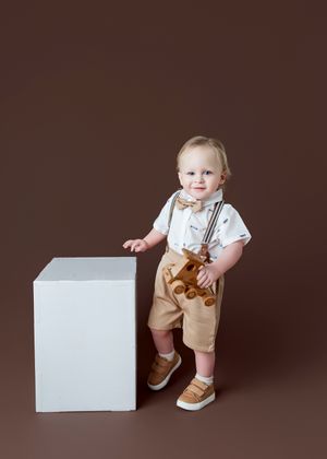 Toddler boy in suspenders, bow tie, and khaki shorts poses beside a white cube on a brown background.