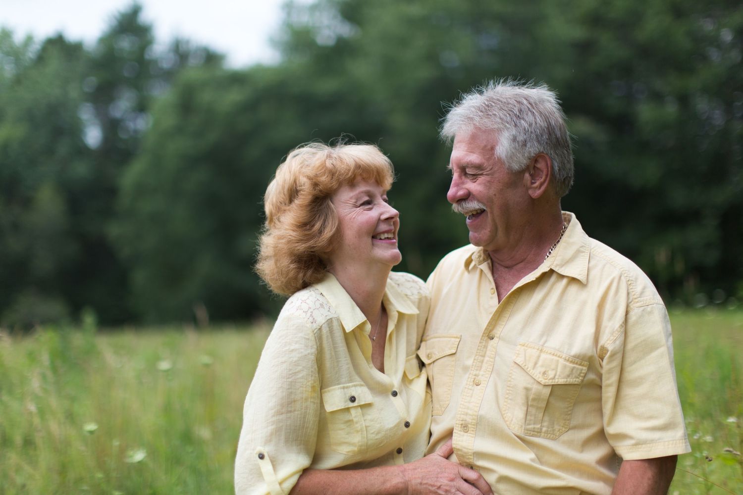 An elderly couple in yellow shirts sitting together and laughing in a sunny outdoor setting.