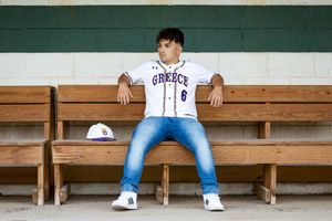 Teen boy in Greece baseball jersey sitting in dugout wearing jeans.