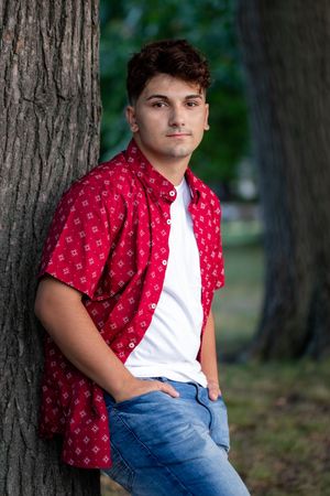 Young man in red patterned shirt and jeans leaning against a tree outdoors.