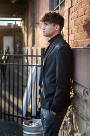 Young man in black blazer leans against brick wall outdoors in urban alley with dramatic lighting.