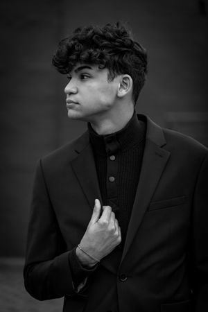 Young man with curly hair in black suit and turtleneck, posing confidently in a dramatic black and white portrait.