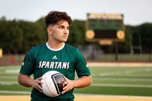 A young male soccer player in a green Spartans jersey holds a ball on a field with a scoreboard in the background.