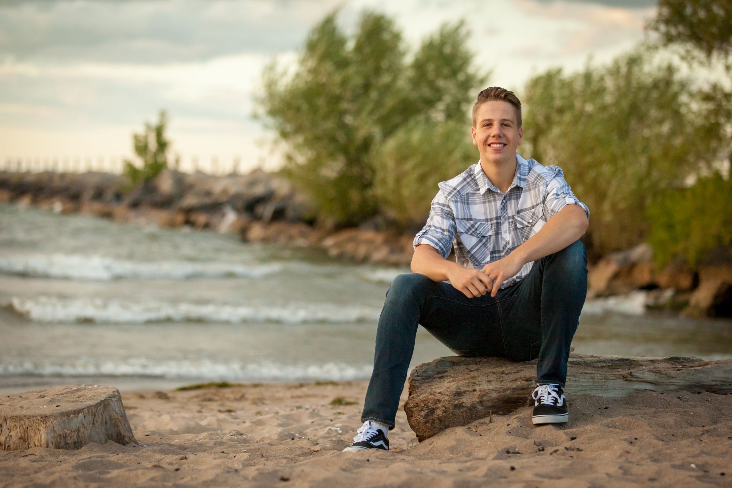 Young man smiling on beach, sitting on driftwood with rocky shoreline and trees in background.