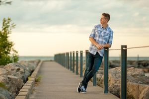 Young man in plaid shirt and jeans leaning on railing of waterfront boardwalk with rocky shoreline at sunset.