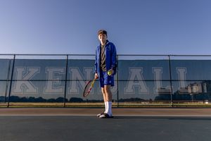 A young male tennis player in blue uniform holds a racket on an outdoor court with Kendall text on fence.