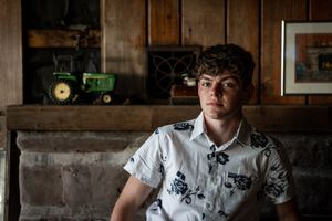Teen boy in floral shirt sits before rustic fireplace with John Deere tractor decor in cozy cabin setting.